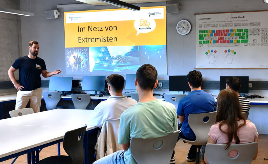 Der Respekt Coach steht in einem Klassenraum vor einer Gruppe Jugendlicher und referiert. Im Hintergrund läuft eine Präsentation mit dem Titel "Im Netz von Extremisten".
