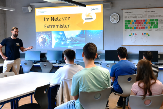 Der Respekt Coach steht in einem Klassenraum vor einer Gruppe Jugendlicher und referiert. Im Hintergrund läuft eine Präsentation mit dem Titel "Im Netz von Extremisten".