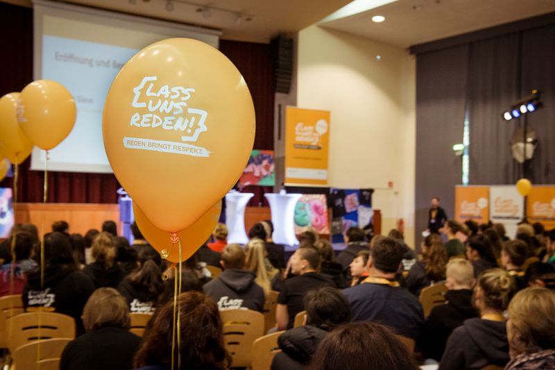 Im Fokus ein orangener Ballon mit dem Programm-Logo "Lass uns reden!". Im Hintergrund Menschen in einem Saal.