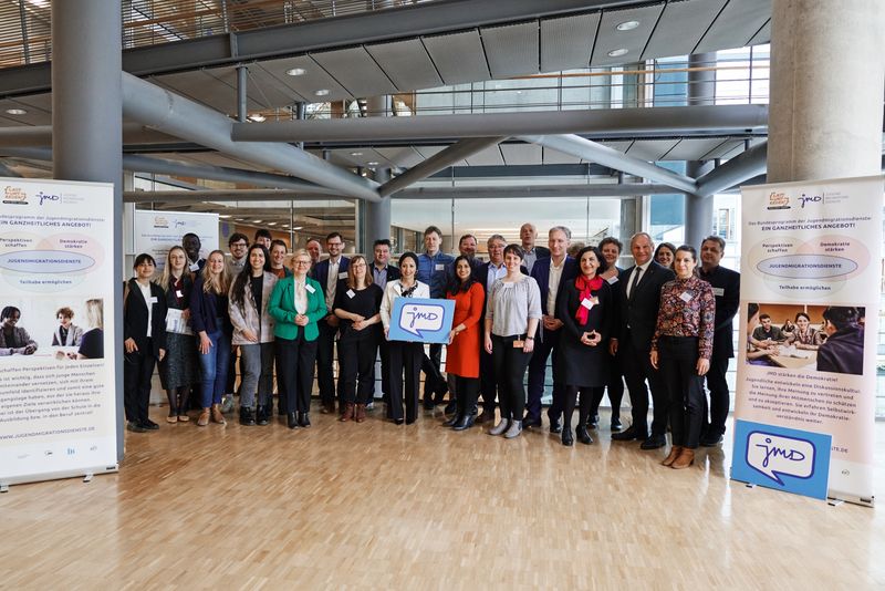 Eine Gruppe Menschen im Parlament steht der Kamera zugewandt. An den Seiten Roll-ups der Jugendmigrationsdienste.