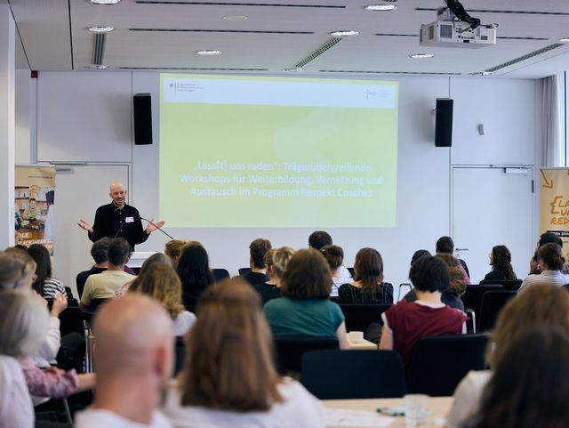 Ein Saal voller Menschen, die von hinten zu sehen sind. Vorne an einem Podium steht ein Mann, der spricht und gestikuliert.
