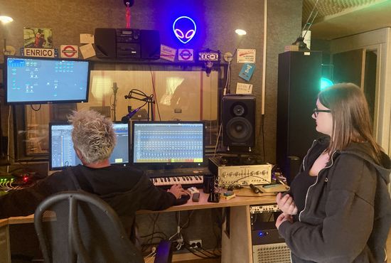 Zwei Personen in einem Tonstudio. Vor ihnen stehen mehrere Bildschirme.