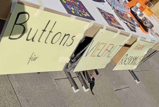 An einem Tisch hängen Schilder, auf denen steht "Buttons für Vielfalt, gegen Rassismus". Auf dem Tisch liegen Postkarten und Stifte und eine Buttonmaschine.