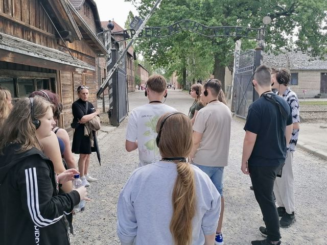 Eine Gruppe steht draußen in einem Gelände und hört einer Person zu.