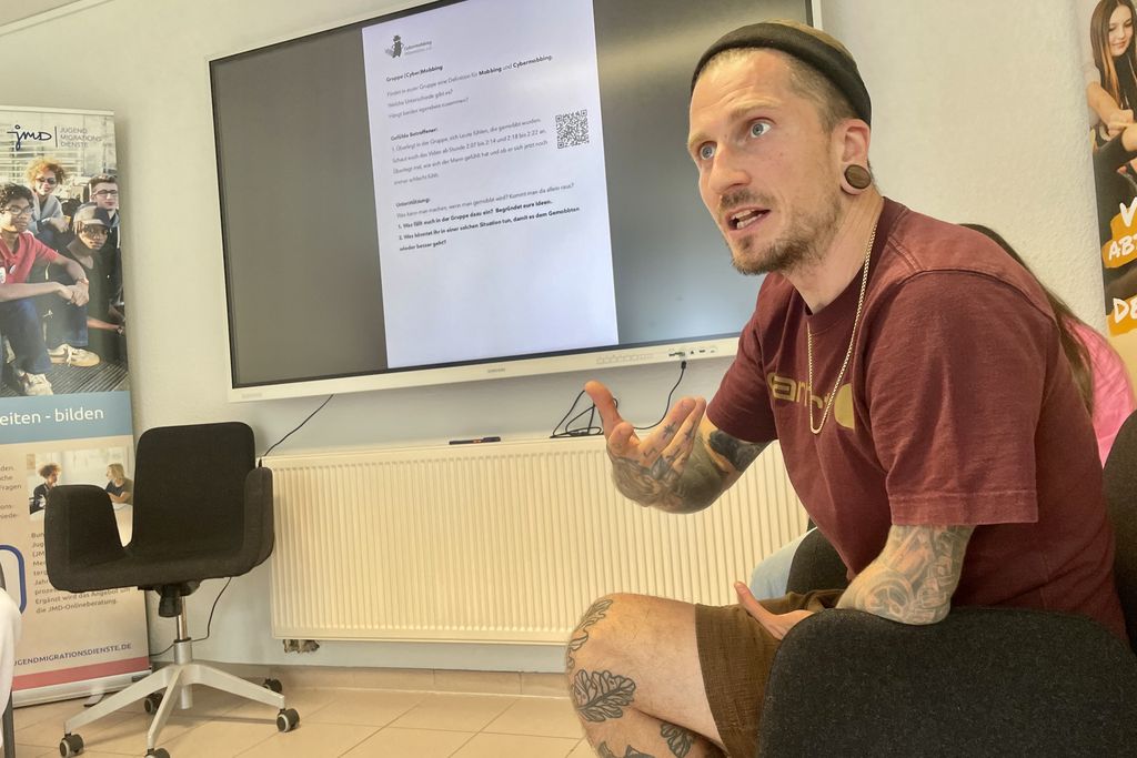 Ein Mann sitzt in einem Seminarraum und gestikuliert. Er schaut nach vorne und spricht.