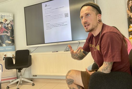 Ein Mann sitzt in einem Seminarraum und gestikuliert. Er schaut nach vorne und spricht.