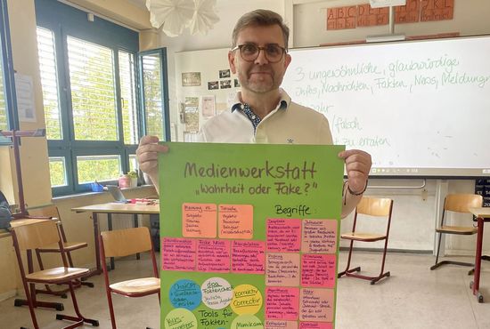 Ein Mann steht in einem Klassenzimmer und hält ein Plakat hoch, auf dem steht: Medienwerkstatt "Wahrheit oder Fake?". Auf dem Plakat sind viele einzelne bunte Post-Its zu sehen.