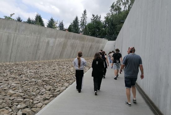 Eine Gruppe, von hinten zu sehen, läuft über einen grauen Boden, rechts und links Betonwände, neben dem Weg liegen graue Steine.