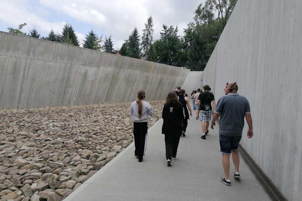 Eine Gruppe, von hinten zu sehen, läuft über einen grauen Boden, rechts und links Betonwände, neben dem Weg liegen graue Steine.