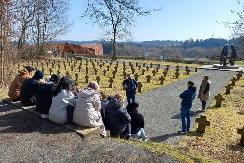 Mehrere junge Personen sitzen mit Blick auf eine Grabstätte.