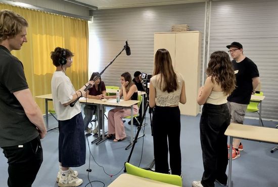 Eine Gruppe Jugendlicher im Klassenzimmer: Eine Person steht an einer Filmkamera, eine andere Person hält ein Mikrofon, zwei Schülerinnen sitzen an einem Tisch und unterhalten sich.