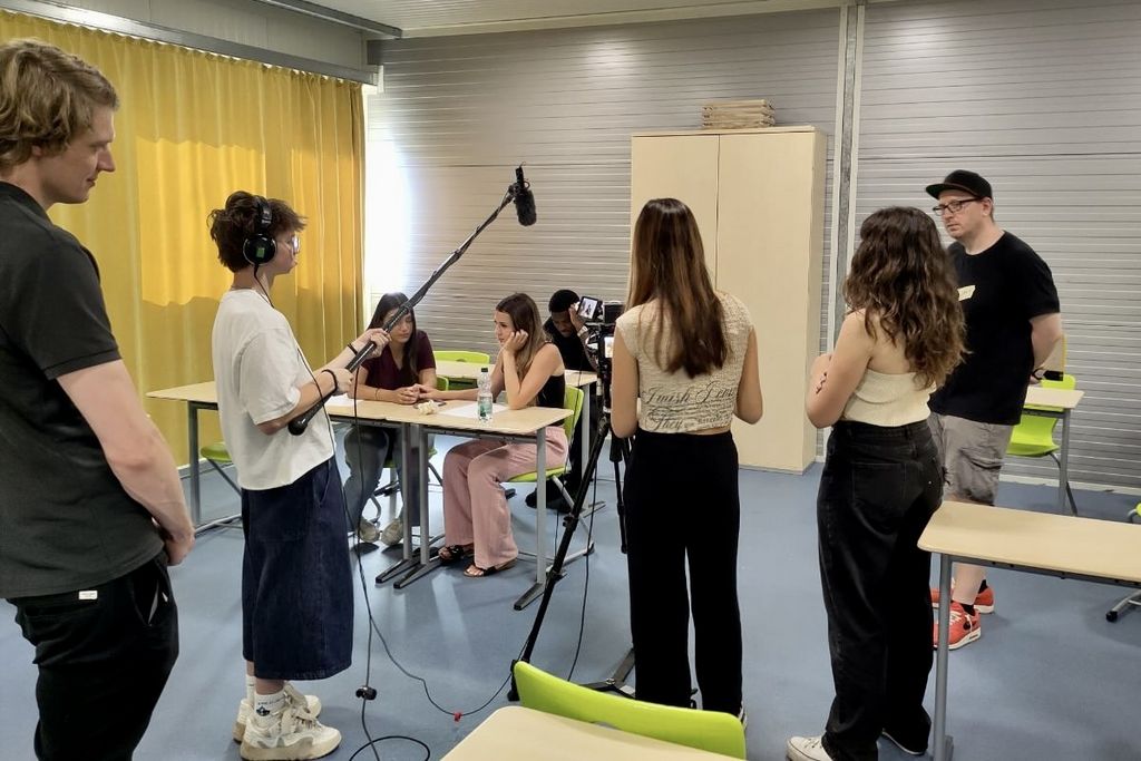 Eine Gruppe Jugendlicher im Klassenzimmer: Eine Person steht an einer Filmkamera, eine andere Person hält ein Mikrofon, zwei Schülerinnen sitzen an einem Tisch und unterhalten sich.