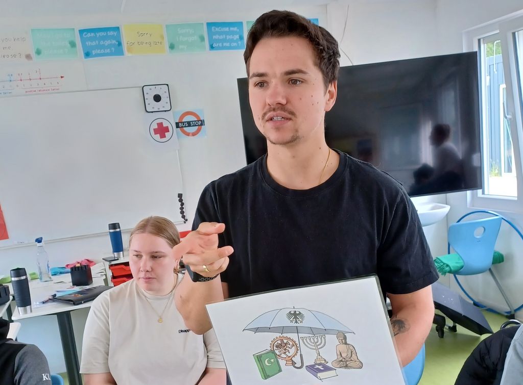 Der Respekt Coach zeigt der Klasse ein Bild mit Symbolen unterschiedlicher Religionen. Das Bild zeigt, dass die Religionsfreiheit durch das Grundgesetz geschützt ist.