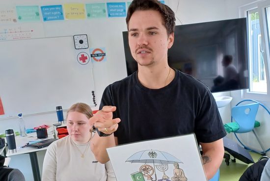 Der Respekt Coach zeigt der Klasse ein Bild mit Symbolen unterschiedlicher Religionen. Das Bild zeigt, dass die Religionsfreiheit durch das Grundgesetz geschützt ist.