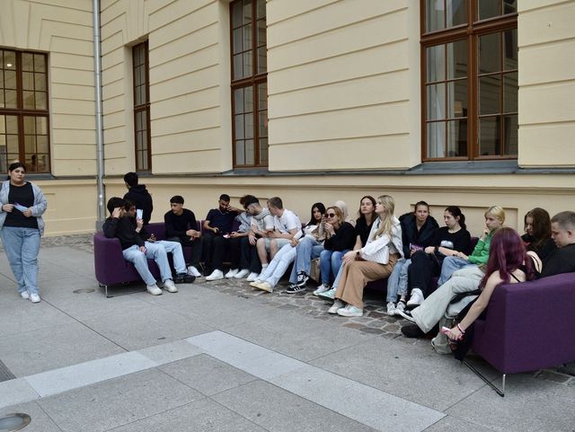 Eine Gruppe junger Menschen sitzt entspannt auf Sofas im Freien vor einem historischen Gebäude mit gelber Fassade.