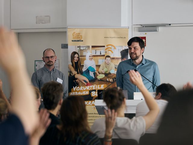 Zwei Männer stehen vor den Menschen im Plenum. Einige Hände sind gehoben.