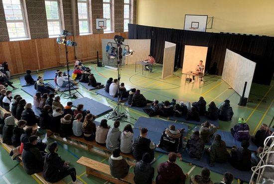 Mehrere junge Personen sitzen auf dem Boden vor zwei vortragenden Personen.