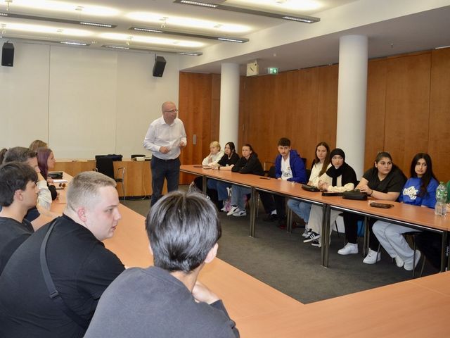Eine große Gruppe von Menschen sitzt in einem Konferenzraum an einem U-förmigen Tisch und hört einem stehenden Mann zu, der spricht. Der Raum hat eine Holzvertäfelung und eine helle Decke mit Beleuchtung.