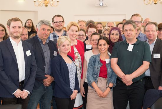 Eine große Gruppe von Menschen steht dicht beieinander und blickt in die Kamera. Viele tragen Namensschilder oder weiße Symbole an der Kleidung. Der Raum ist hell und festlich beleuchtet.
