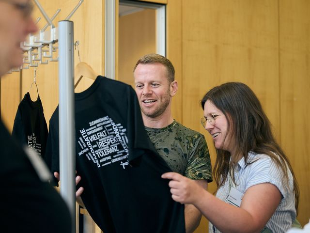 Zwei Personen schauen sich ein schwarzes T-Shirt an, das an einem Kleiderbügel hängt. Auf dem T-Shirt ist eine Wortwolke mit Begriffen wie Respekt und Vielfalt zu sehen.