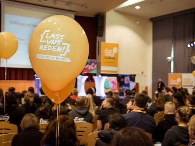Publikum verfolgt eine Bühnenveranstaltung mit Podiumsdiskussion, orangefarbene Luftballons mit der Aufschrift "Lass uns reden!" im Vordergrund.
