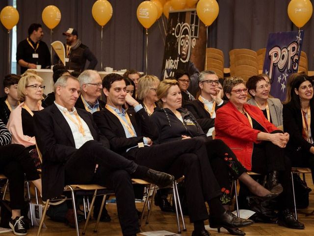Zuschauerinnen und Zuschauer sitzen in einem Veranstaltungsraum und hören aufmerksam zu; gelbe Luftballons schmücken den Hintergrund.