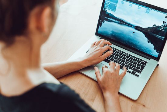 Eine Person sitzt an einem Tisch und arbeitet an einem Laptop. Auf dem Bildschirm ist eine Landschaft mit Wasser und Bergen zu sehen.