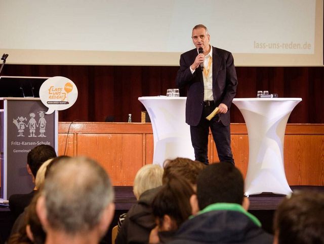 Ein Mann spricht auf einer Bühne vor Publikum, neben ihm stehen zwei weiße Stehtische und ein Rednerpult mit Veranstaltungslogo.