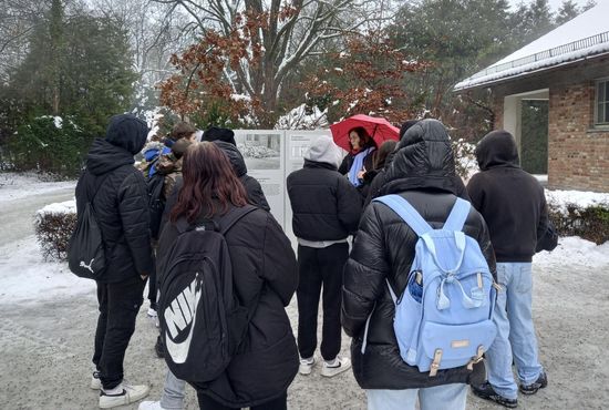 Eine Gruppe von Menschen in Winterkleidung steht im Freien vor einer Gedenktafel. Der Boden ist schneebedeckt, und im Hintergrund sind Bäume und ein Gebäude zu sehen.