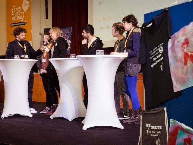 Sechs Personen diskutieren auf einer Bühne an Stehtischen; rechts sind kreative Ausstellungsstücke wie T-Shirts und Bilder zu sehen.