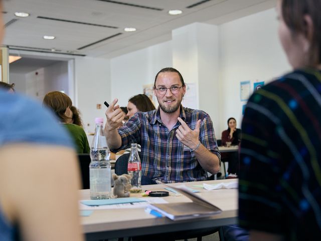 Eine Gruppe Menschen an einem Tisch. Ein Mann, der in der Runde sitzt, spricht und gestikuliert zu den anderen.