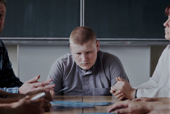 Ein junger Mann mit ernstem Blick sitzt an einem Tisch, während mehrere Personen um ihn herum gestikulieren. Im Hintergrund ist eine Tafel zu sehen.