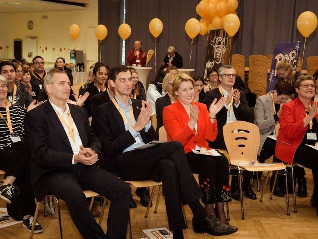Menschen sitzen in einem Veranstaltungsraum, applaudieren und feiern ein Ereignis; orangefarbene Luftballons schmücken den Hintergrund.