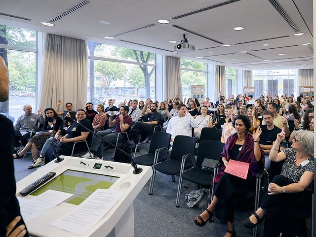 Viele Menschen sitzen in einem Plenum in mehreren Stuhlreihen in einem großen Saal und schauen nach vorne, wo jemand am Podium spricht.