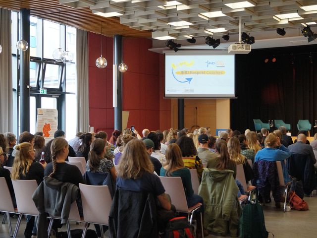 Teinehmende sitzen in Stuhlreihen und blicken Richtung Bühne und Präsentation.