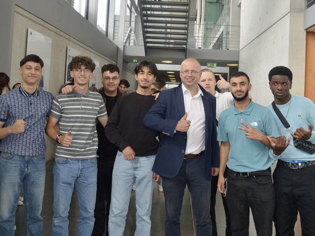 Eine Gruppe junger Männer steht zusammen mit einem Mann im Anzug in einem modernen Gebäude mit Glasfassade. Sie lächeln in die Kamera und zeigen Handgesten wie Daumen hoch.