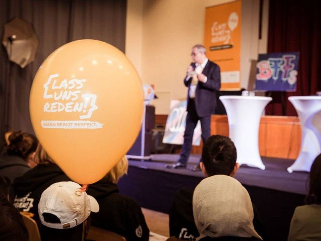 Ein Redner spricht auf einer Bühne vor Publikum; im Vordergrund ein großer orangefarbener Ballon mit der Aufschrift "Lass uns reden!".