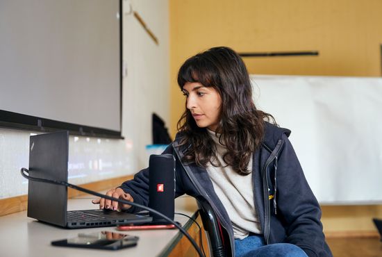 Eine junge Frau sitzt an einem Tisch und tippt mit einer Hand auf einen aufgeklappten Laptop vor sich. Sie schaut auf den Bildschirm des Laptops. Im Hintergrund erkennt man eine Leinwand und einen Flipchart. Der Hintergrund lässt vermuten, dass sich die Frau in einem Klassenzimmer befindet. Auf dem Hoodie der Frau ist ein Teil des Logos des JMD-Programms Respekt Coaches zu sehen.
