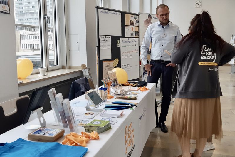 Zwei Menschen unterhalten sich an einem Tisch mit Materialien in einem Foyer.