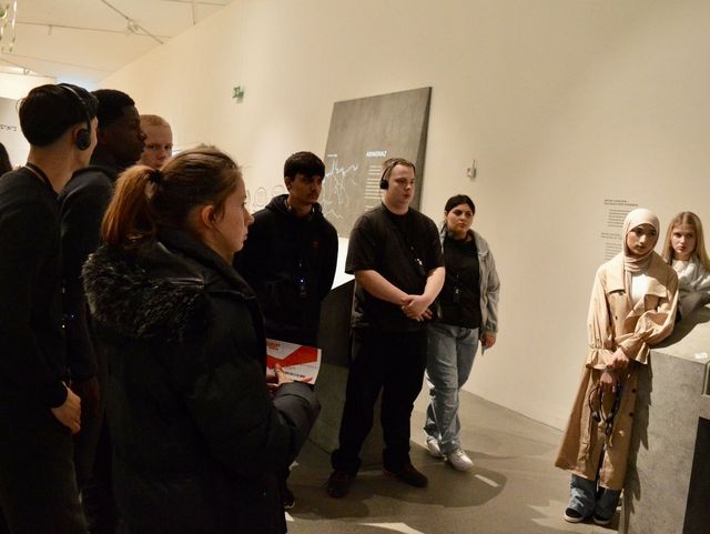 Eine Gruppe von Menschen steht in einem Ausstellungsraum mit weißen Wänden und hört einer Person zu, die etwas erklärt. Einige Besucher tragen Kopfhörer, andere halten Broschüren oder Notizen in der Hand.