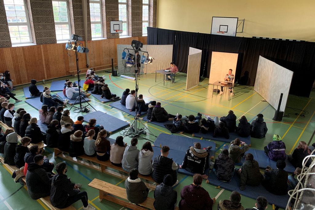 Mehrere junge Personen sitzen auf dem Boden vor zwei vortragenden Personen.