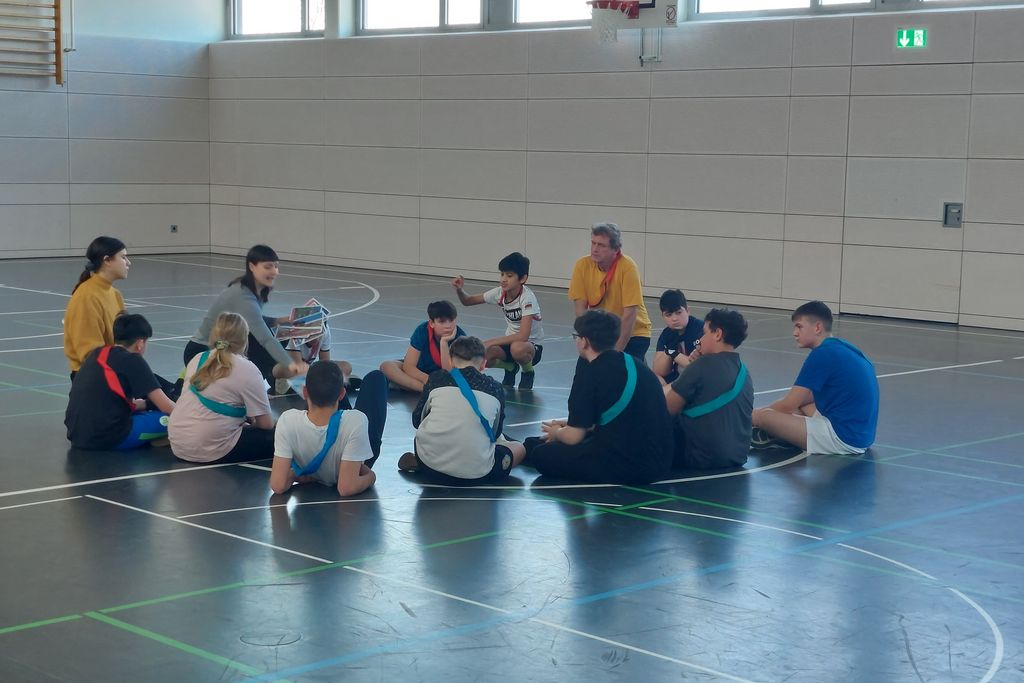 Mehrere junge Personen sitzen auf dem Boden in einer Turnhalle.