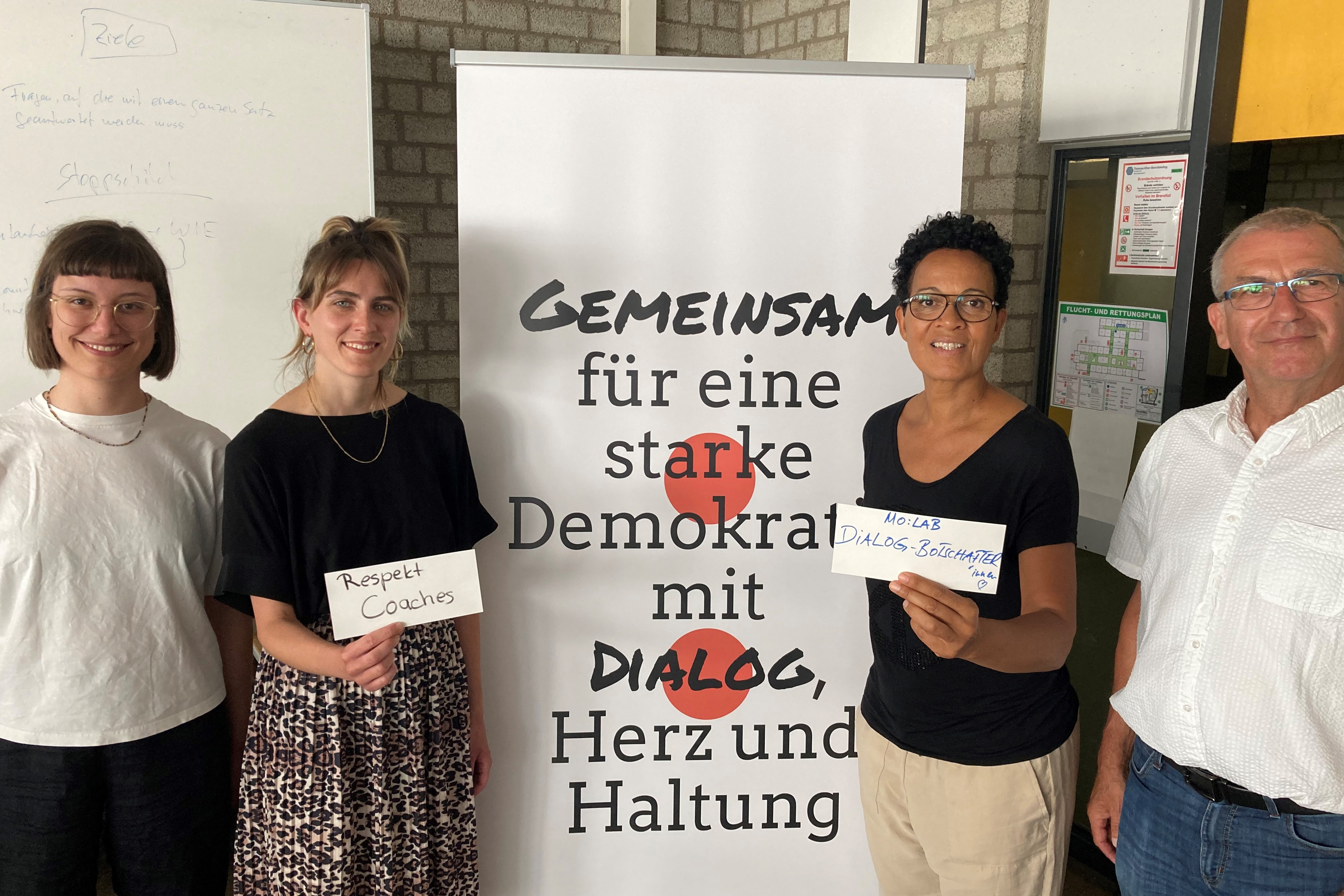 Vier Personen stehen an einem Banner mit dem Text: Gemeinsam für eine starke Demokratie mit Dialog, Herz und Haltung.