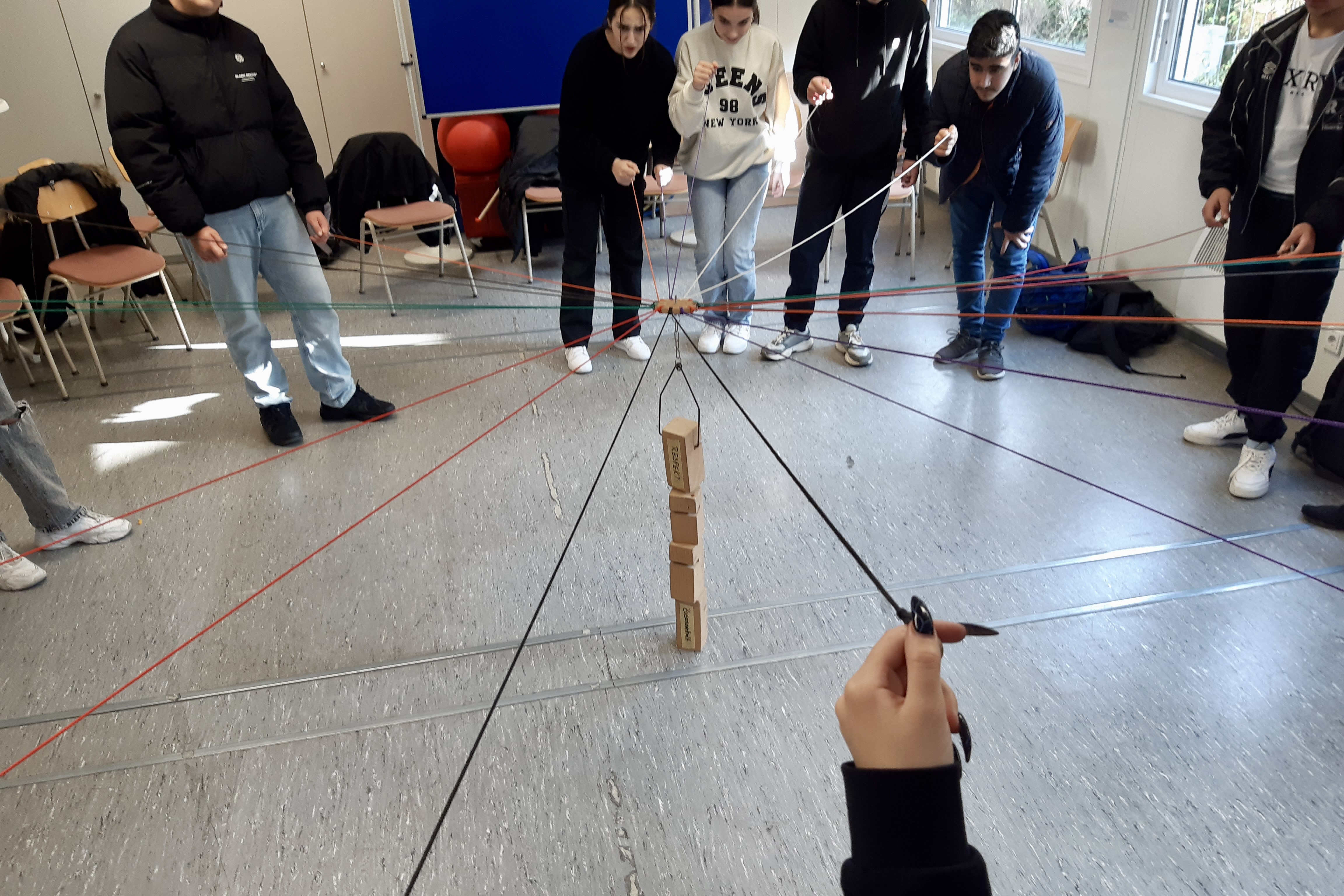 Mehrere junge Menschen stehen in einem Kreis und haben Schnüre in der Hand. Die Schnüre sind mit Holzklötzen in der Mitte der Gruppe verbunden. Über die Schnüre werden die Holzklötze zu einem Turn gestapelt.