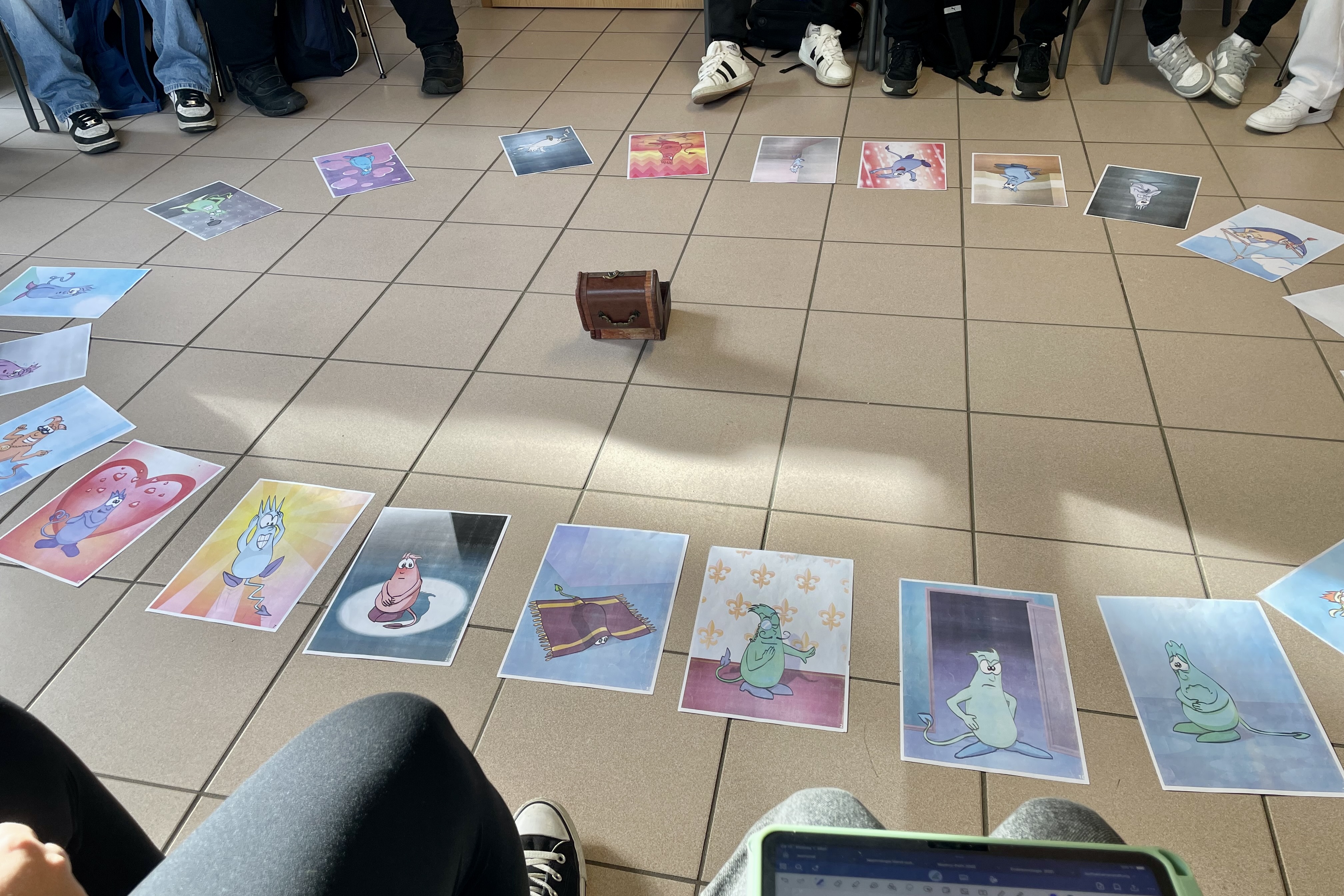 Auf dem Boden liegen in einem Kreis Bilder von Abbildungen mit einem Monster, das auf jedem Blatt unterschiedliche Gefühle darstellt. Um die Blätter herum sind Stühle und Personen zu erkennen.
