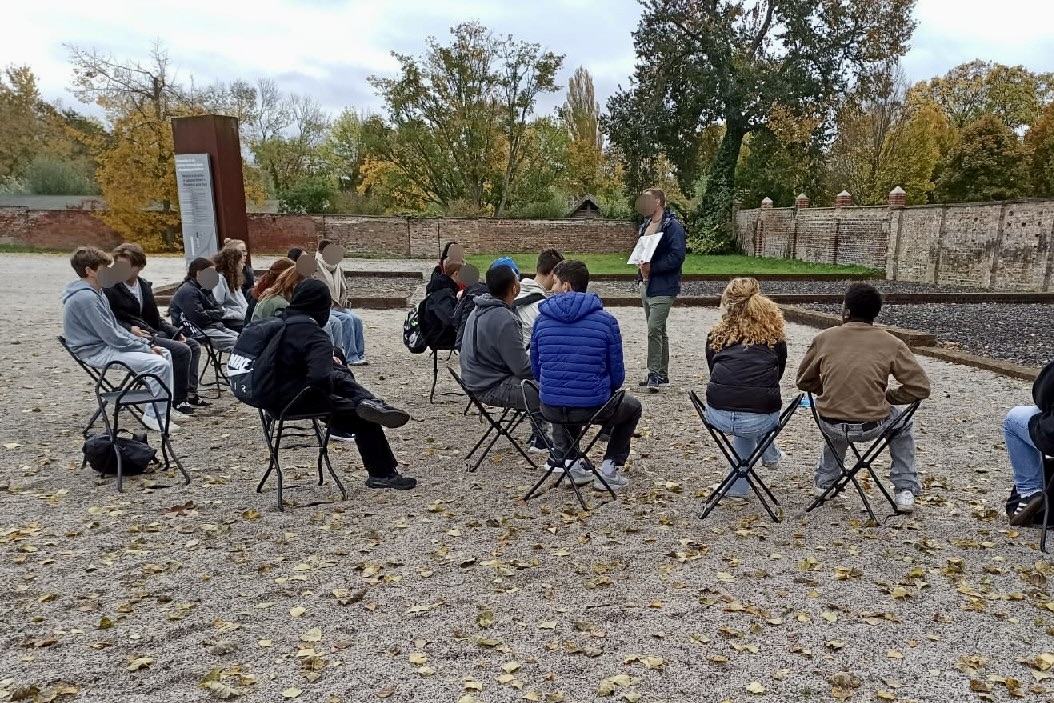 Eine Gruppe junger Menschen auf einem Außengelände einer Gedenkstätte. Vor der Gruppe steht eine erwachsene Person und zeigt etwas auf einem Papier.