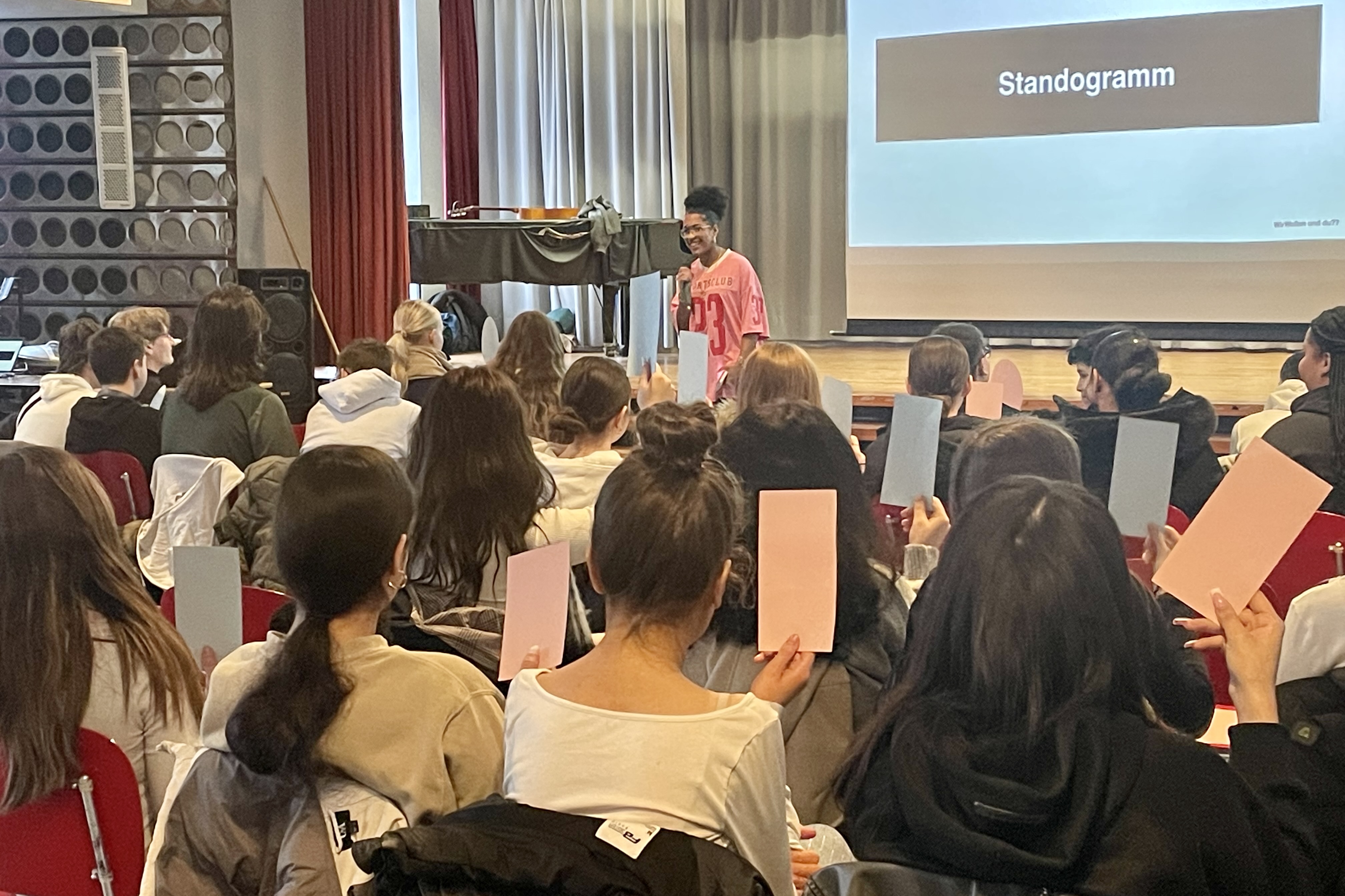 Eine große Gruppe junger Menschen ist von hinten zu sehen. Sie sitzen in einer Aula. Eine Person mit Mikrofon steht vor der Gruppe. Im Hintergrund wird an eine Leinwand das Wort "Standogramm" gebeamt.