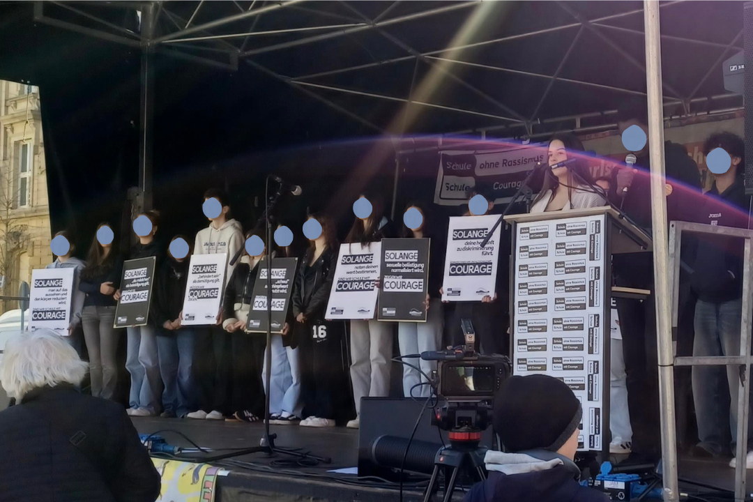 Eine Reihe junger Menschen auf einer Bühne. Sie haben Schilder vor sich. Eine junge Frau am Podium spricht ins Mikro.
